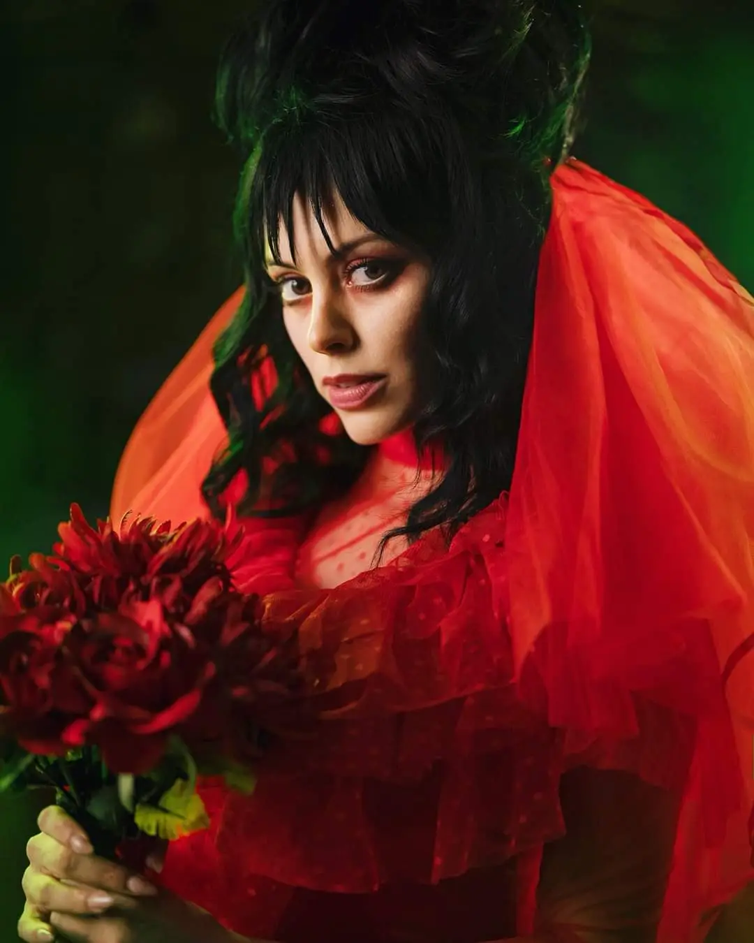 A woman with dark hair and dramatic makeup, wearing a red ruffled dress and veil, holds a bouquet of red roses while looking…