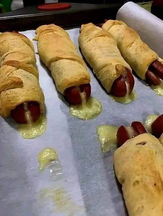 A close-up photo shows several baked hot dogs wrapped in golden-brown pastry, with melted yellow cheese visibly oozing out…