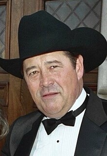 A man wearing a black cowboy hat, a black tuxedo with a white shirt and black bow tie, looks directly at the camera with a…