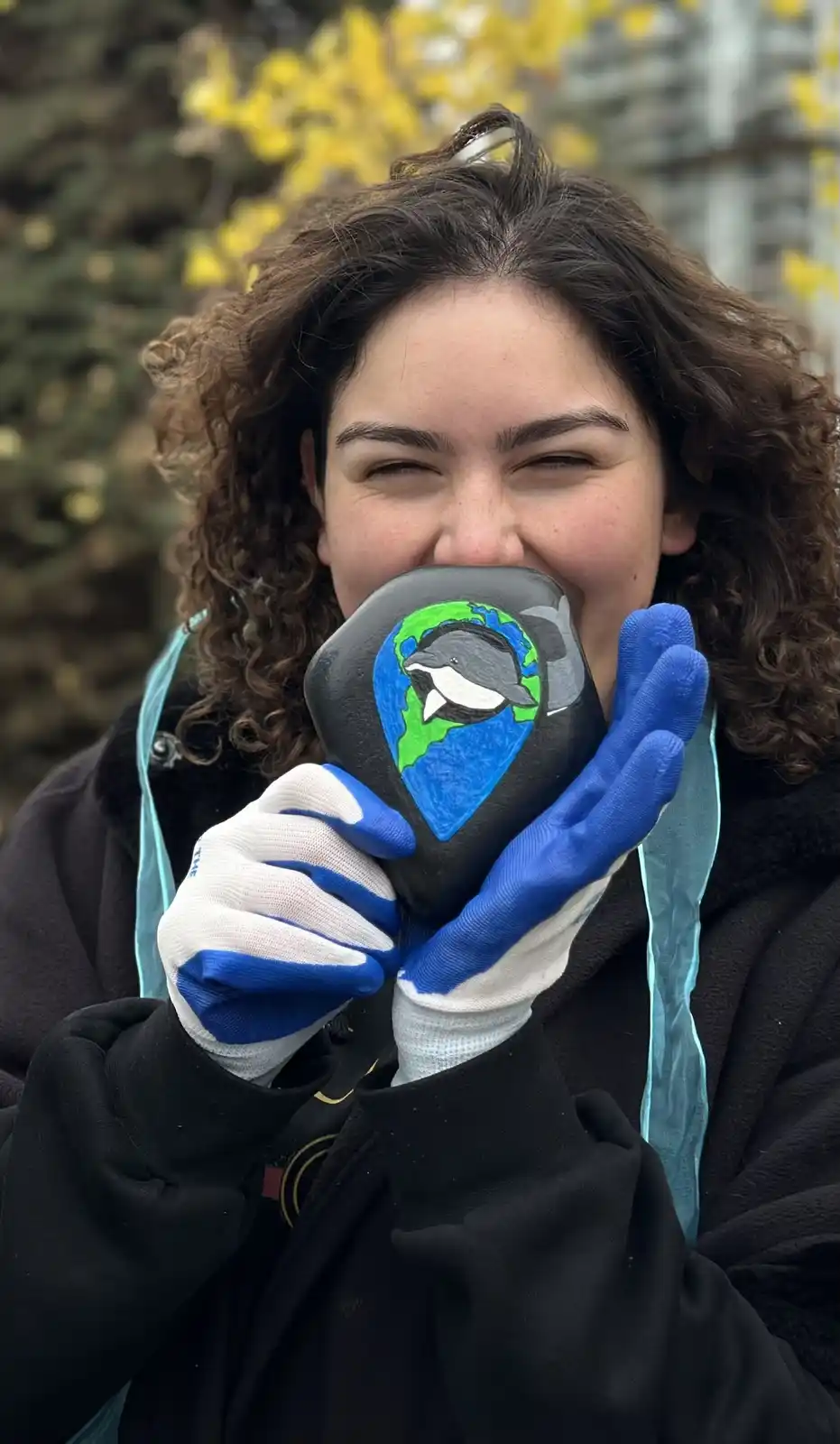 A person with curly brown hair and wearing blue and white gloves holds a painted rock in front of their face. The rock…
