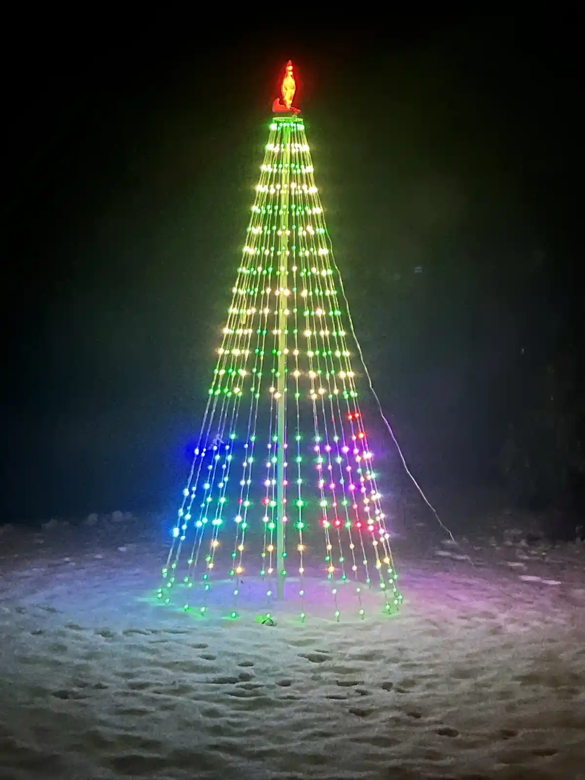 A tall, cone-shaped Christmas tree made of vertical strings of multicolored lights stands in snow at night, topped with a…