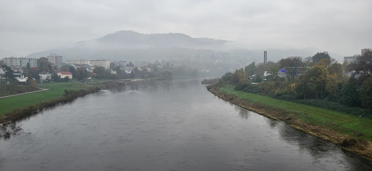 A wide river flows through a misty, overcast landscape with towns on both banks and hills in the background. The overlaid…