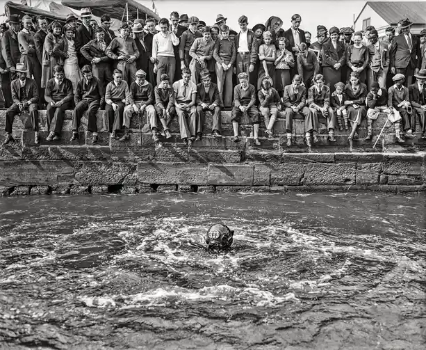 a black-and-white historical photo shows a crowd of people, mostly children and teens in period clothing, sitting and…