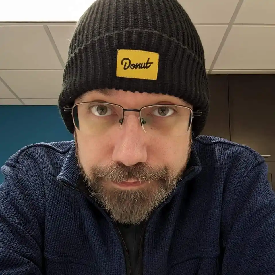 A man with glasses and a beard wears a black knit beanie featuring a yellow rectangular patch with the word 'Donut' in black…