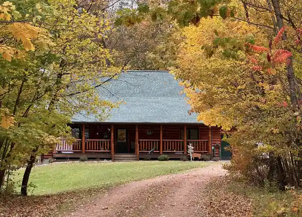 A picturesque log cabin surrounded by autumn foliage with yellow and orange leaves, a gravel path leads to the porch…