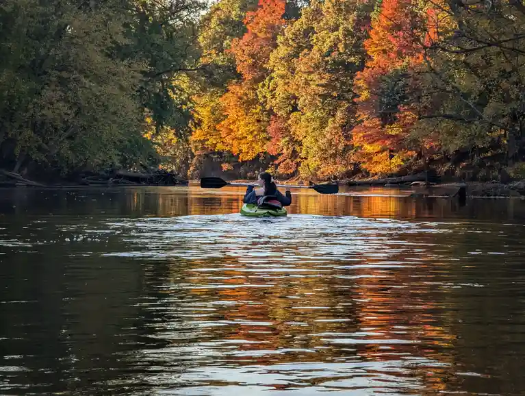 A person in a green kayak paddles down a calm river surrounded by vibrant autumn foliage reflecting in the water. The person…