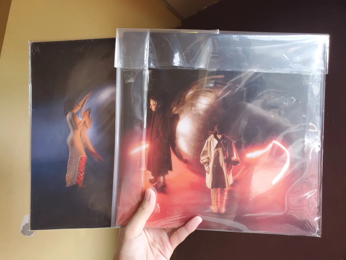 A hand holds two sealed vinyl record sleeves side-by-side; the left sleeve shows two figures in a blue-lit scene, while the…