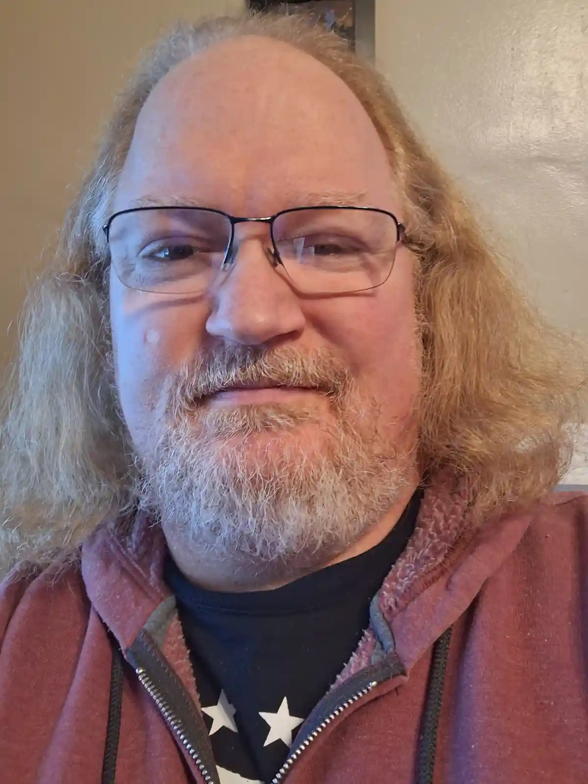A close-up photo of a middle-aged man with long, wavy, reddish-blonde hair, a graying beard, and glasses, wearing a maroon…