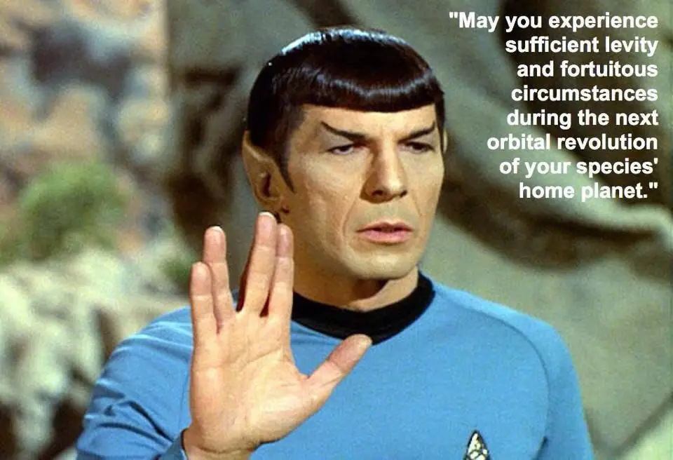 A still image of the character Spock from Star Trek, wearing a blue uniform and raising his hand in the Vulcan salute. White…