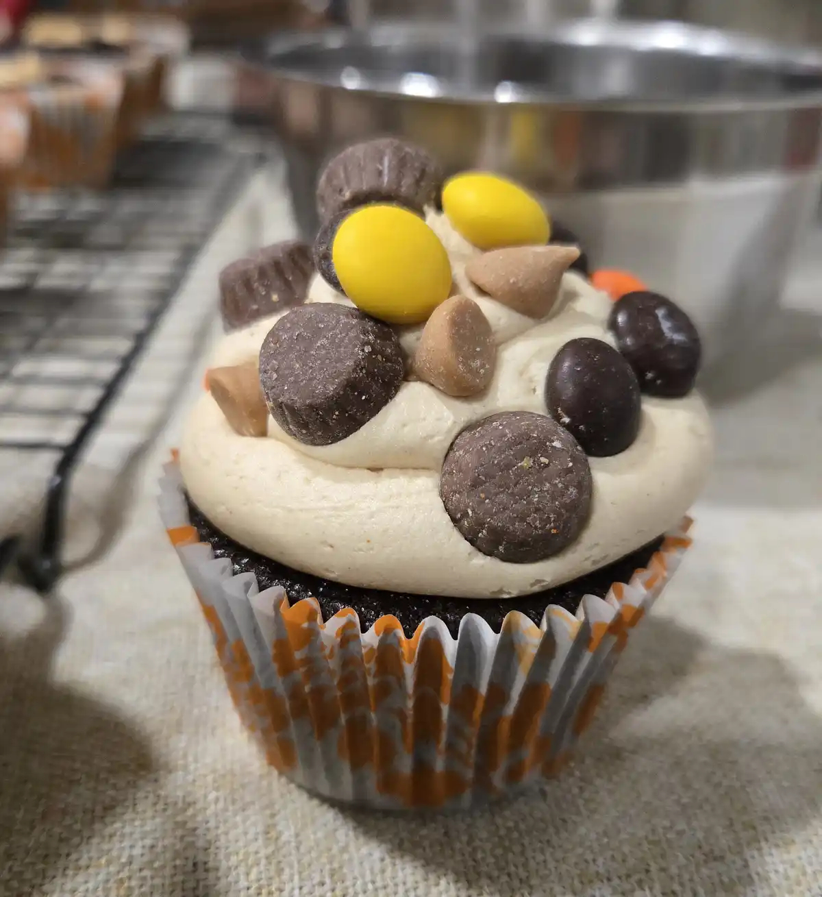 A close-up photo of a chocolate cupcake with light-colored frosting, decorated with an assortment of chocolate candies…