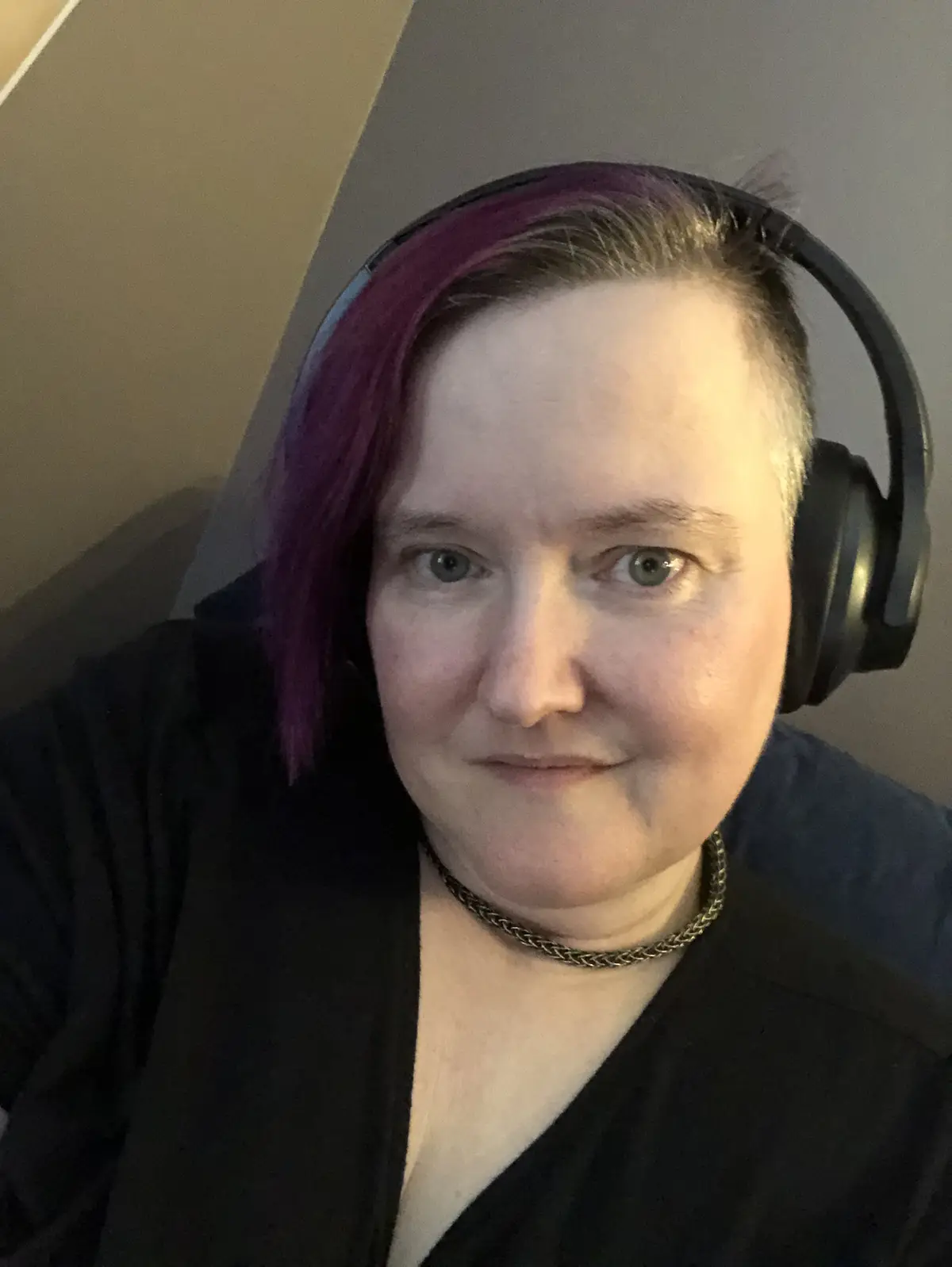 A person with short hair dyed purple on one side and gray on the other wears black over-ear headphones and a black top with…