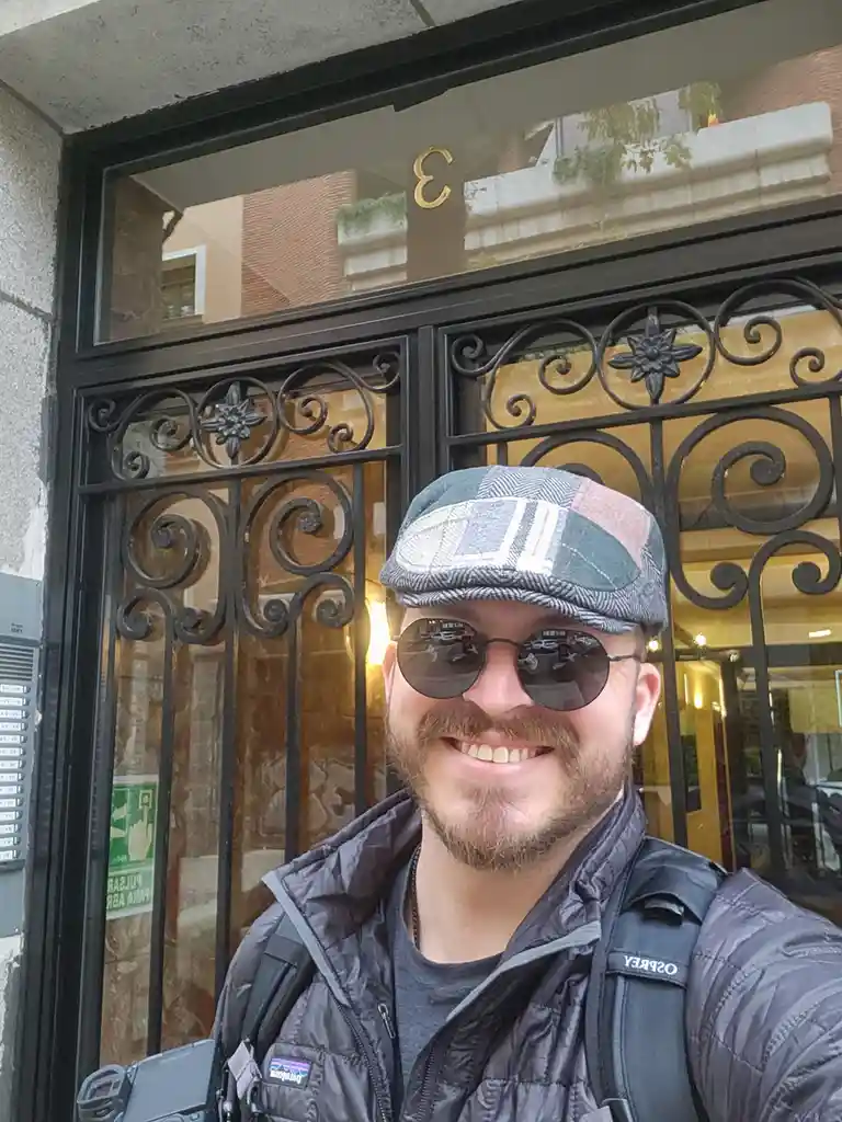 A smiling man with a beard, sunglasses, and a plaid cap takes a selfie in front of an ornate black wrought-iron gate. Above…