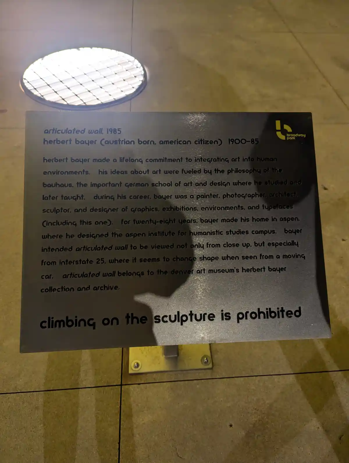 A close-up of a dark metal plaque mounted on a tiled sidewalk, describing Herbert Bayer's 1985 sculpture 'Articulated Wall'…