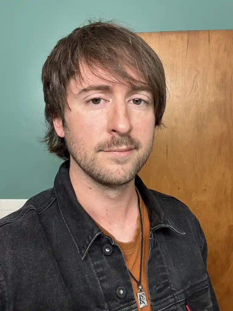 A man with brown hair and a mustache, wearing a dark denim jacket over an orange shirt, looks directly at the camera with a…