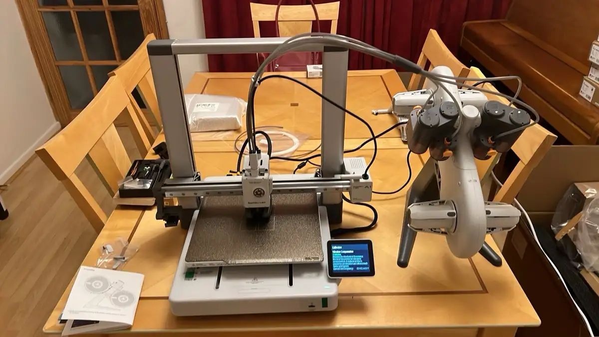 A 3D printer sits on a wooden dining table, next to a white robotic arm with multiple joints and cables. The printer's small…