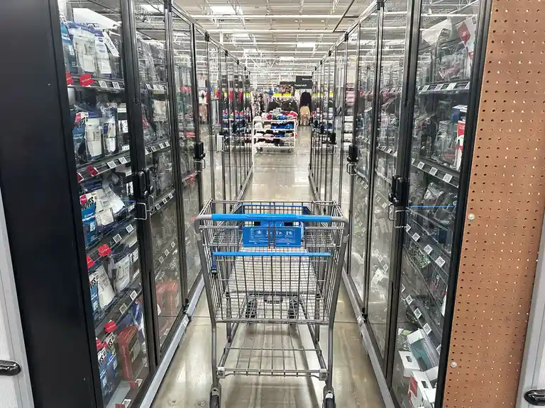 An empty Walmart shopping cart sits in the middle of a long aisle flanked by tall glass-fronted refrigerated cases filled…