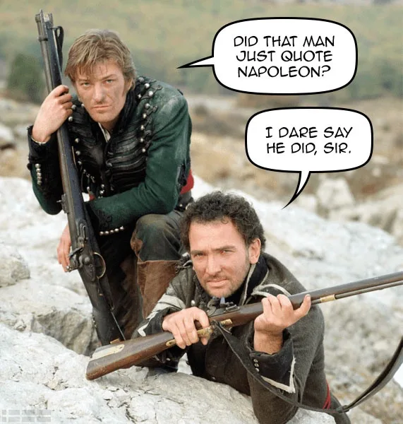 Two men in historical military attire crouch on rocky terrain, each holding a musket. The man in the background has a stern…