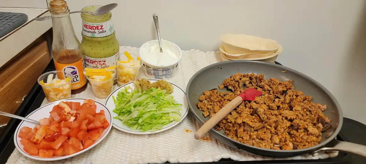 A kitchen counter displays ingredients for tacos: a frying pan with cooked meat and mushrooms, plates of chopped tomatoes…