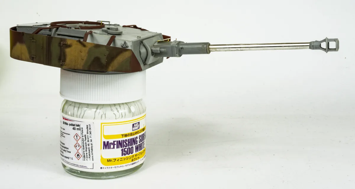 A scale model of a tank with camouflage paint is mounted on top of a bottle of Mr. Hobby white surface finishing thinner…