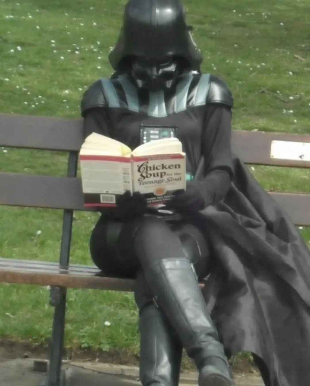 A person dressed in a full Darth Vader costume, including helmet, cape, and boots, is seated on a park bench reading an open…