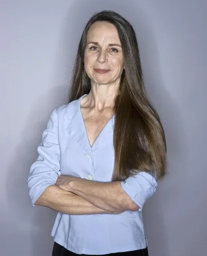 A woman with long brown hair, wearing a light blue button-up shirt, stands with her arms crossed against a plain gray…