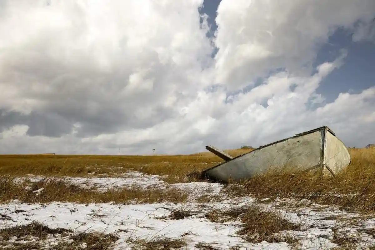 A weathered, white wooden boat lies abandoned in a field of dry grass and patches of snow under a cloudy sky. The boat is…