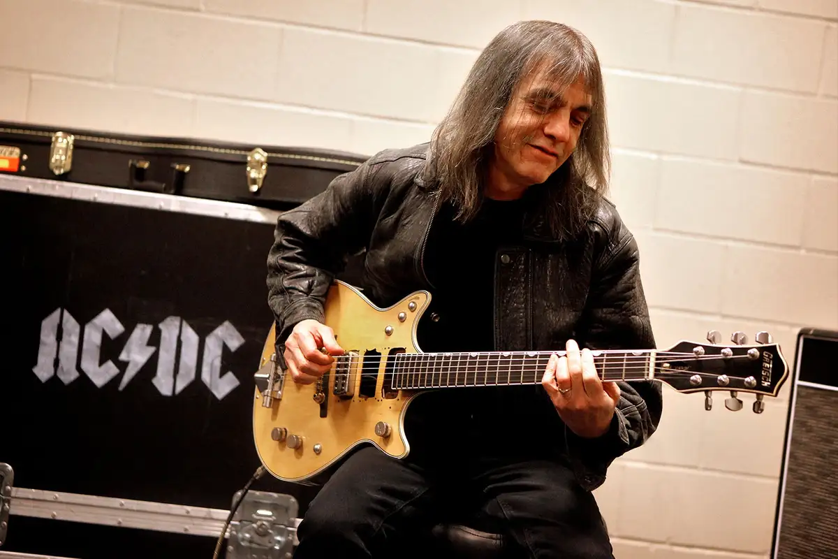 a man with long gray hair wearing a black leather jacket sits playing a light-colored Gretsch electric guitar. Behind him is…
