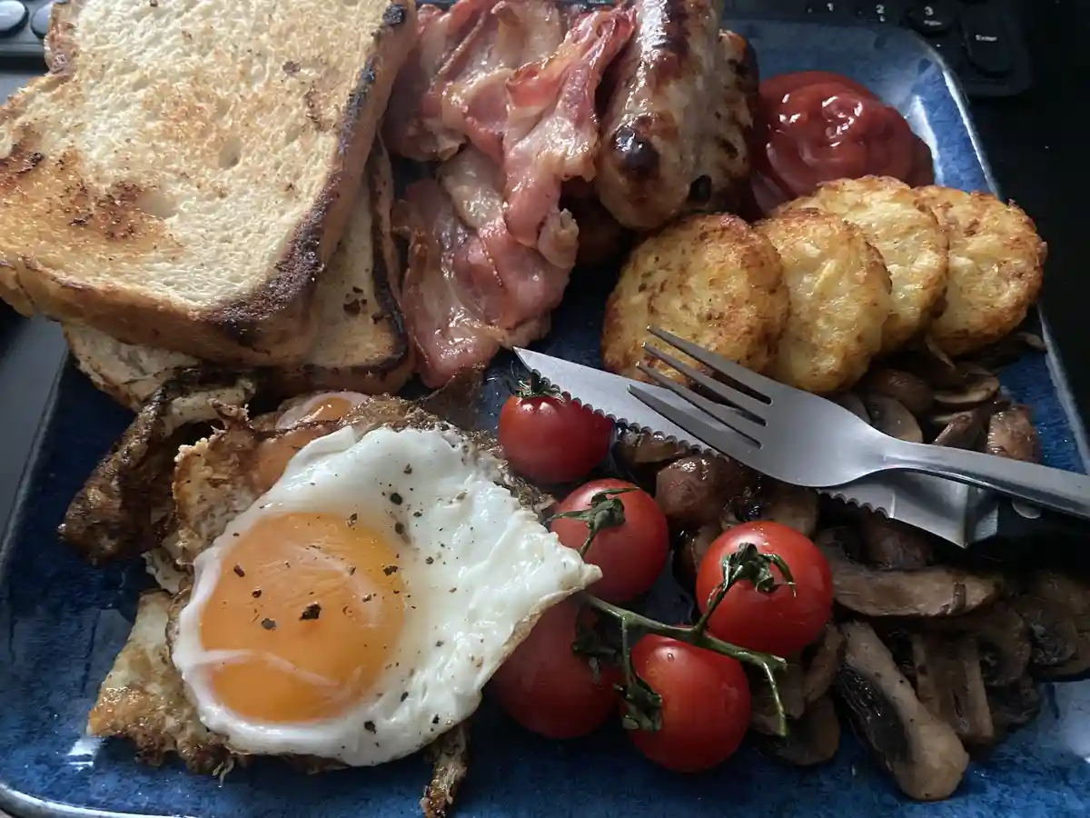 A close-up photo of a full English breakfast on a blue plate, featuring fried egg, bacon, sausage, toast, hash browns…