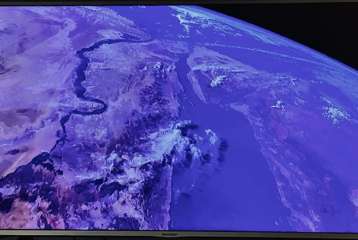 A television screen displays a satellite image of Earth, showing a winding dark river cutting through purple-hued land and…