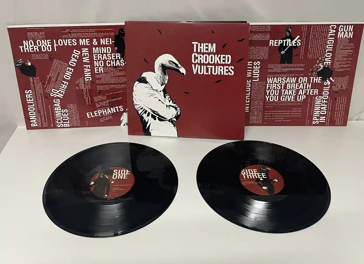 Three red album covers for 'Them Crooked Vultures' are displayed behind two black vinyl records. The central cover features…