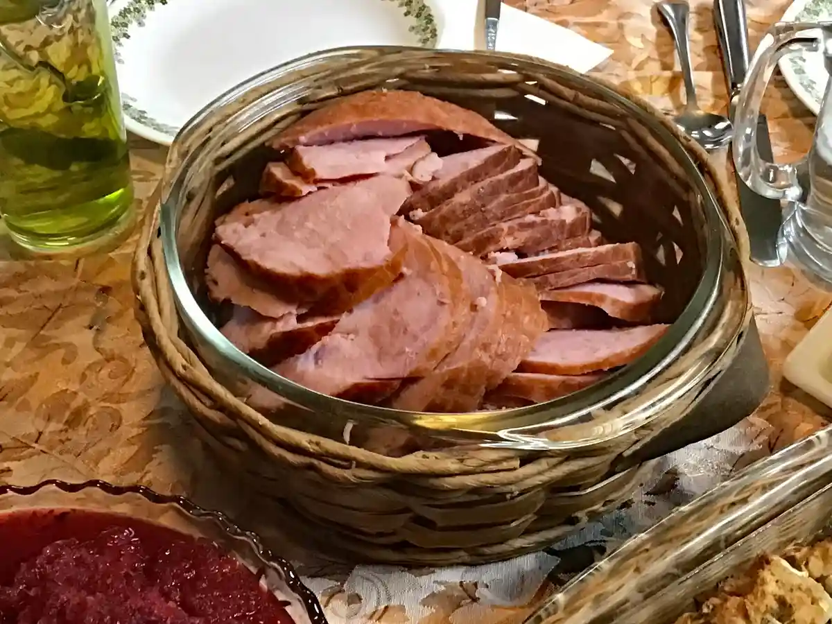 A woven basket filled with sliced ham sits on a patterned tablecloth, surrounded by dining utensils, a glass of green…