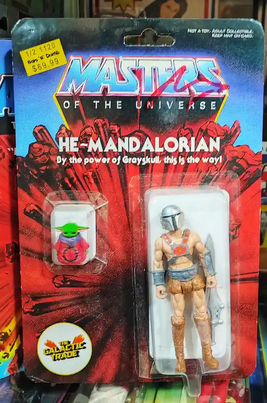 A Masters of the Universe action figure package features a Mandalorian character with a silver helmet and armor, holding a…