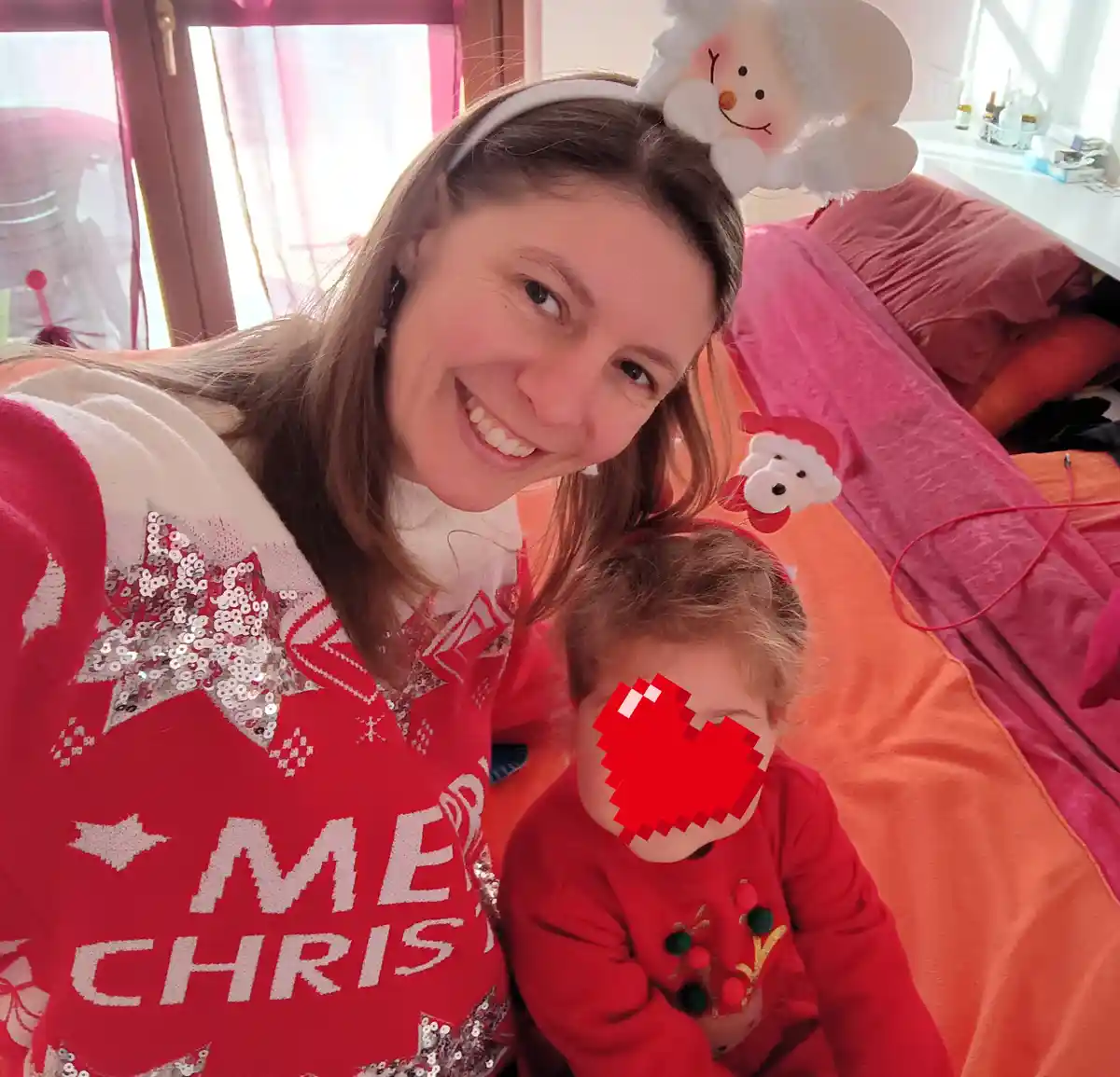 A woman in a red 'Merry Christmas' sweater with sequined stars smiles while wearing a snowman headband, posing with a child…