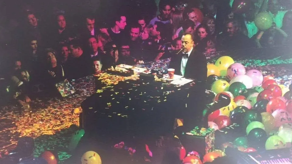 A man in a tuxedo sits at a piano surrounded by confetti and balloons, with a crowd watching him. The text overlaid on the…
