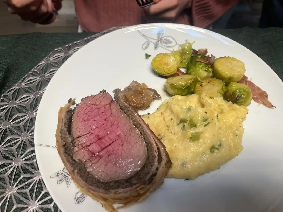 A close-up photo shows a plated beef wellington, sliced to reveal a pink, medium-rare center, served with creamy mashed…