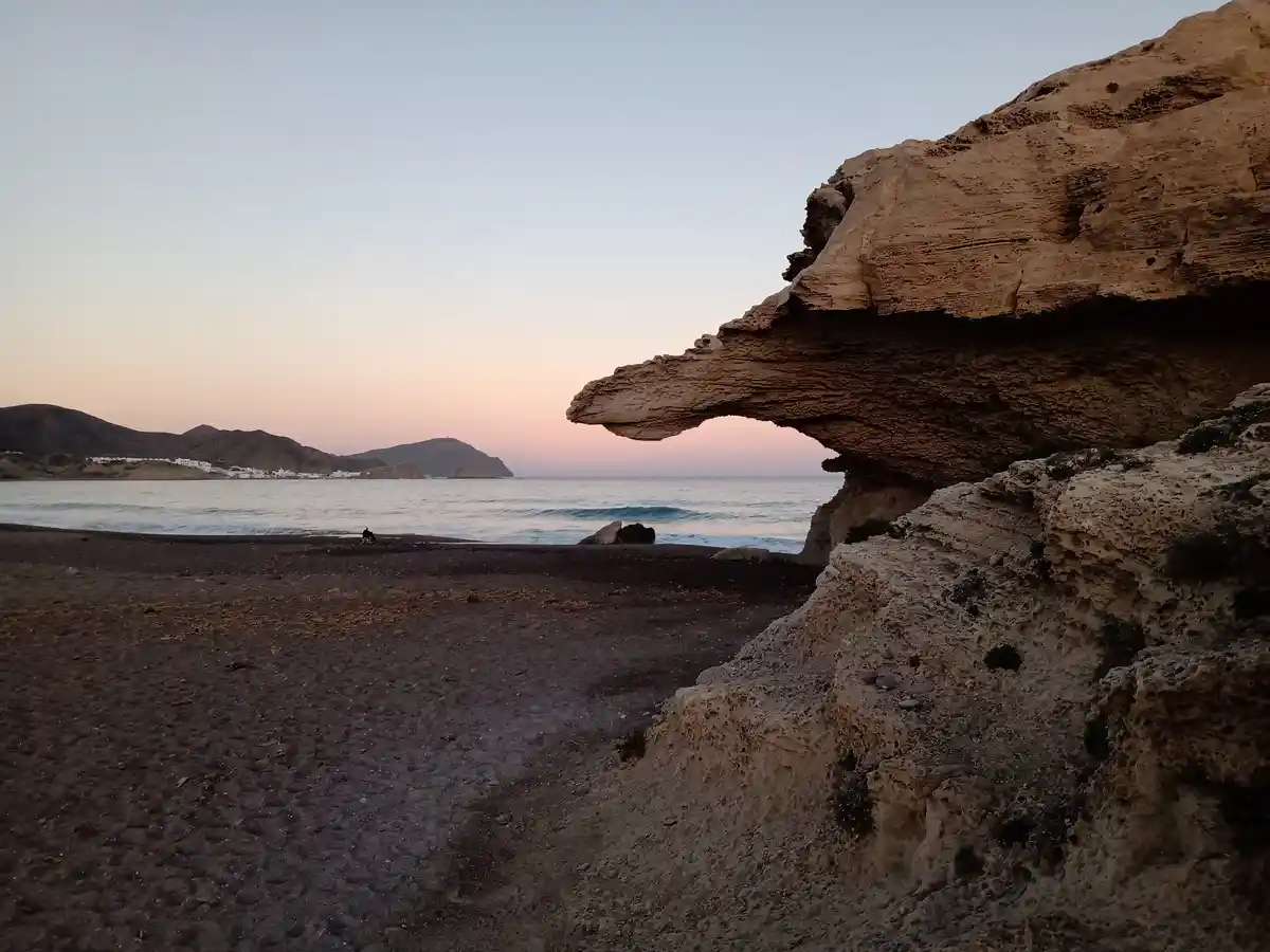 A serene beach scene at dusk with a large, overhanging rock formation on the right, dark sand in the foreground, gentle…