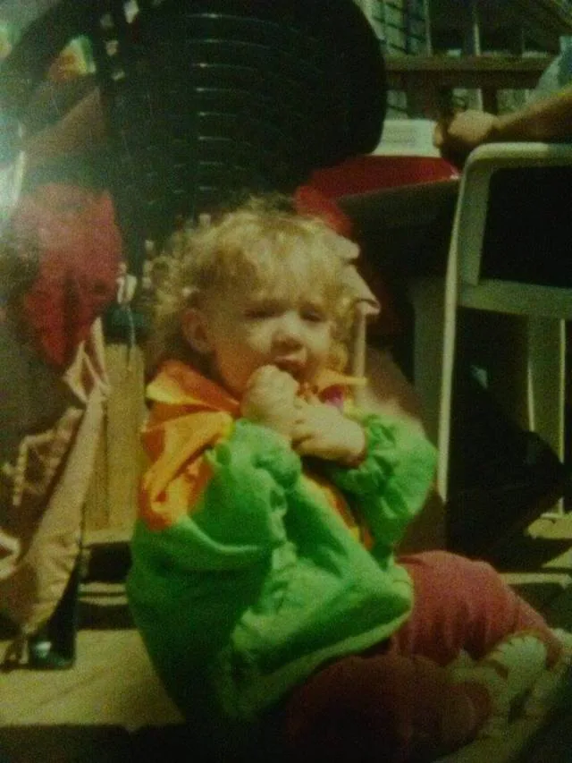 A grainy, low-resolution photo of a young child with curly blonde hair, wearing a green and orange jacket and red pants…