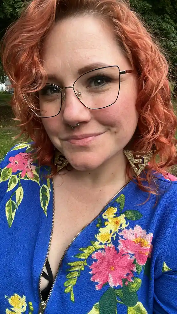 A woman with curly reddish hair, wearing black-rimmed glasses, a nose piercing, and large gold triangle earrings, smiles…