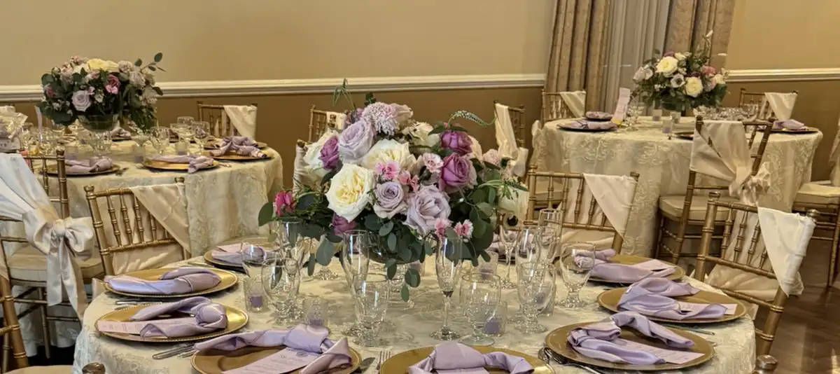 A formal banquet hall is set with round tables covered in white lace tablecloths, gold chiavari chairs with white sashes…