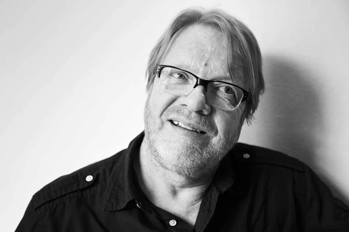 A black-and-white portrait of a middle-aged man with glasses and a beard, smiling while looking slightly upward. He wears a…