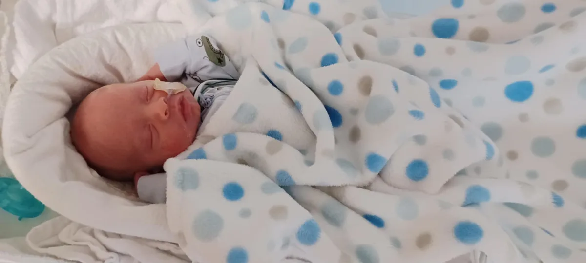 A newborn baby is sleeping peacefully in a hospital bed, wrapped in a white blanket with blue and gray polka dots. The…