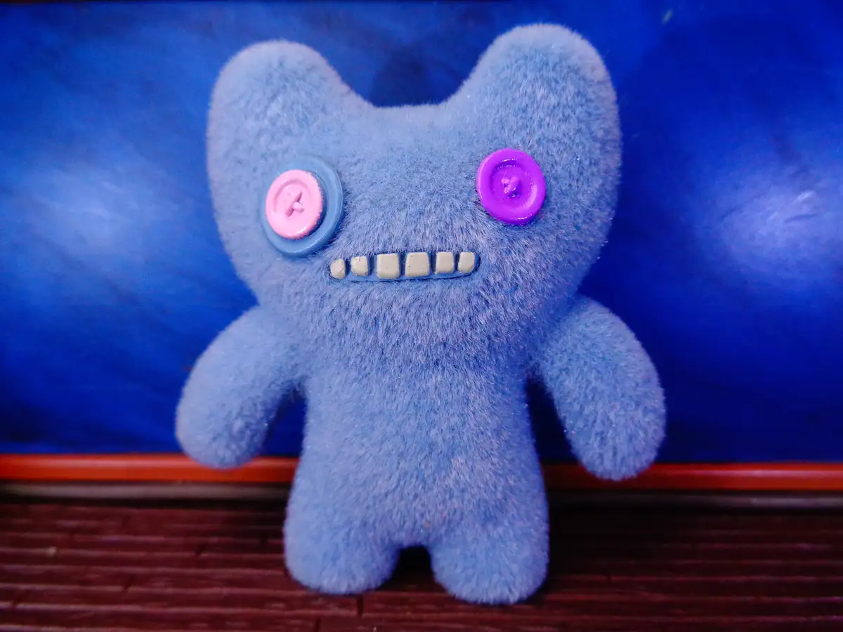 A blue, fuzzy plush toy with mismatched button eyes (one pink, one purple) and a row of small, square teeth, standing…
