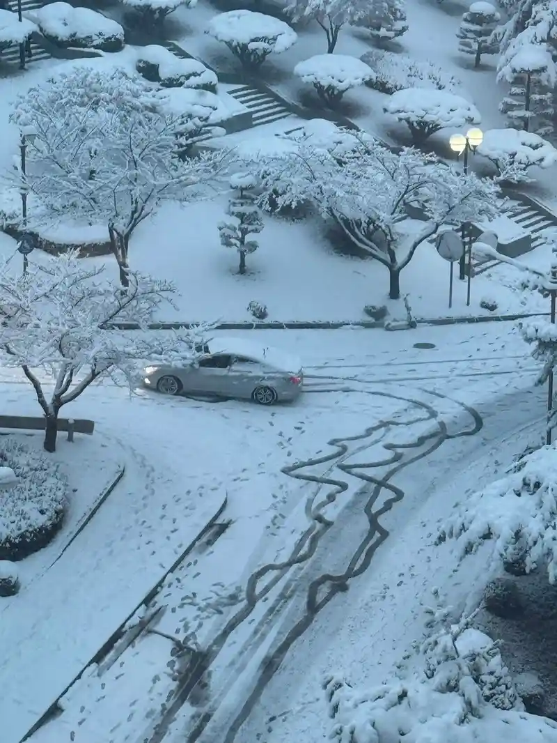 An aerial view shows a silver car stuck in a snow-covered parking lot, making wide, looping tire tracks in the snow as it…