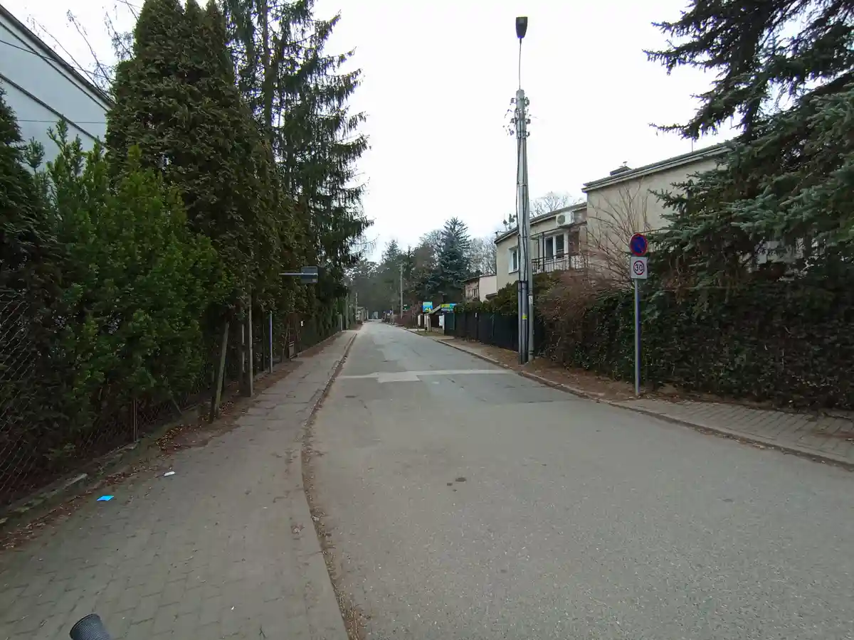 A quiet residential street with sidewalks, trees, and houses under an overcast sky. A traffic signpost on the right displays…