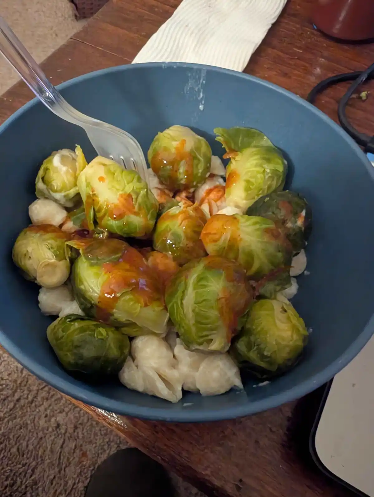 A blue bowl contains cooked brussels sprouts and gnocchi mixed together, drizzled with a reddish-orange sauce, with a clear…