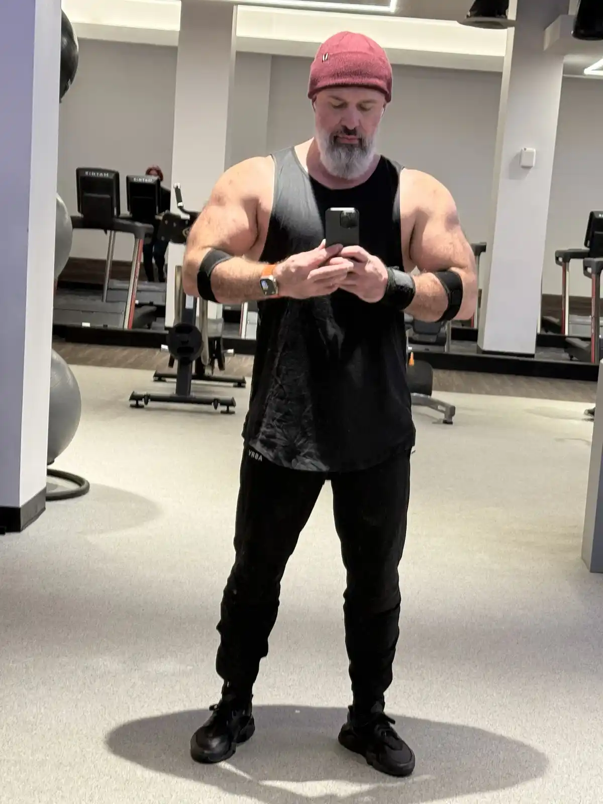 A muscular man with a grey beard and red beanie takes a mirror selfie in a gym, wearing a black tank top and black pants…