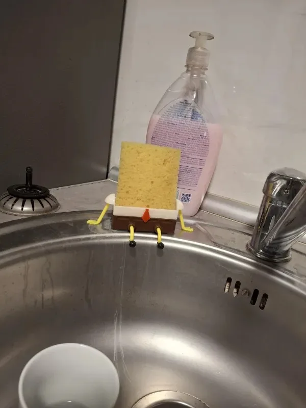 A yellow kitchen sponge is positioned upright on a stainless steel sink edge, with small yellow pipe cleaners attached to…