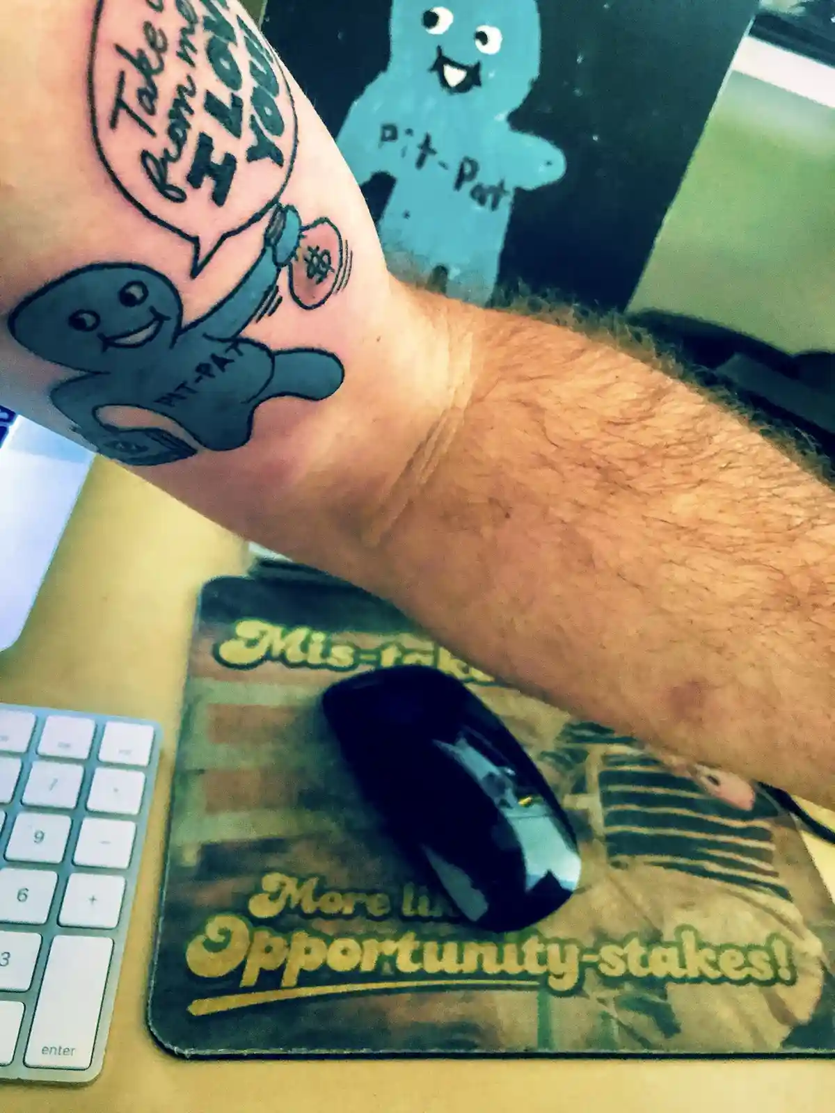 A person's forearm displays a tattoo of a blue, smiling, cartoonish figure labeled 'PIT-PAT' holding a syringe, with a…