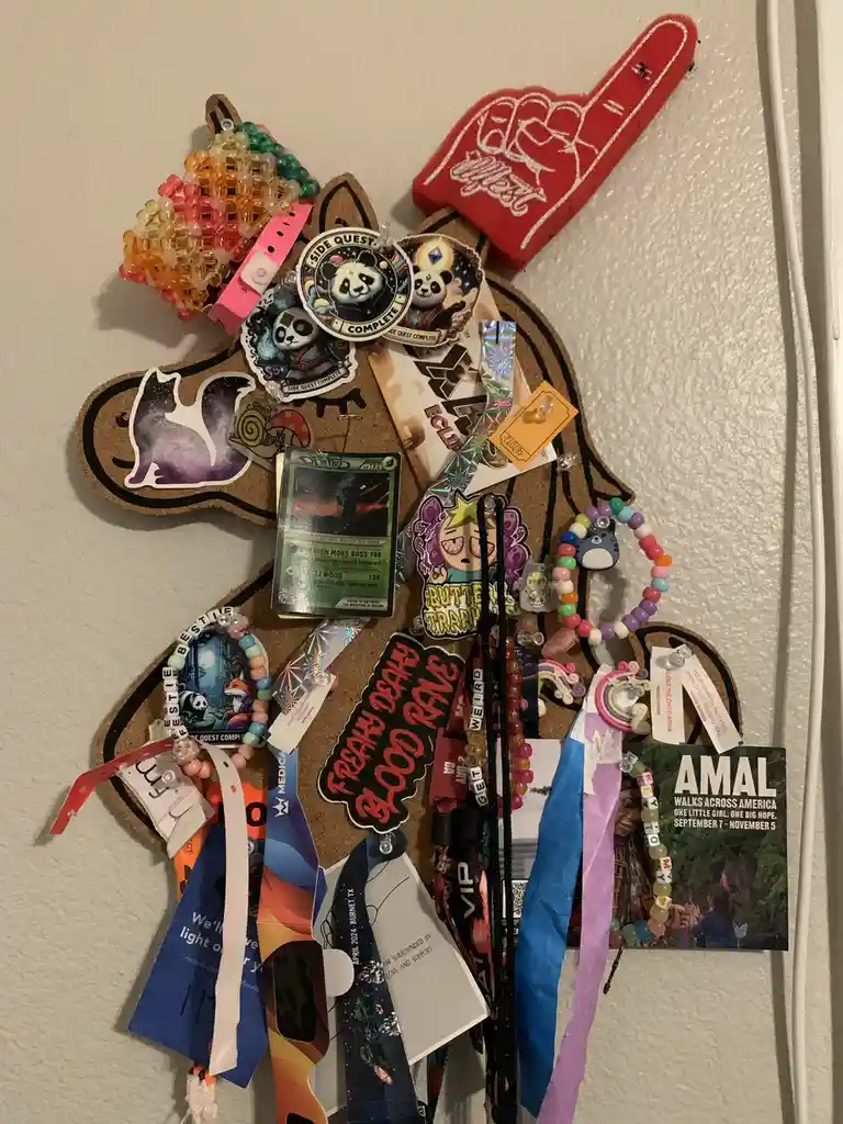 A corkboard shaped like a cartoon girl's head is densely covered with various colorful memorabilia including lanyards…