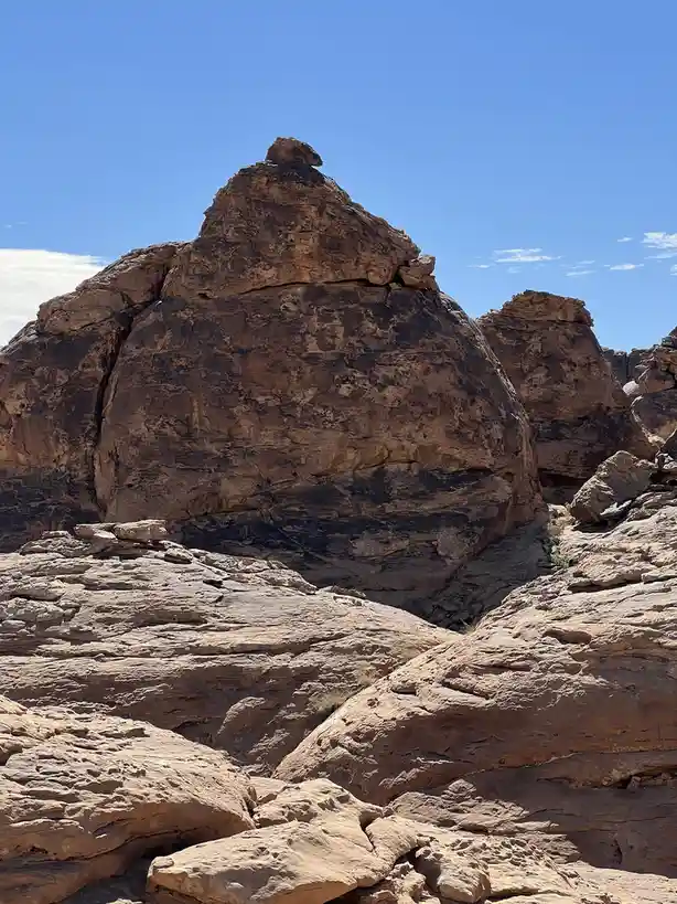 A photograph of a large, textured rock formation under a clear blue sky, with a smaller rock balanced precariously on top…